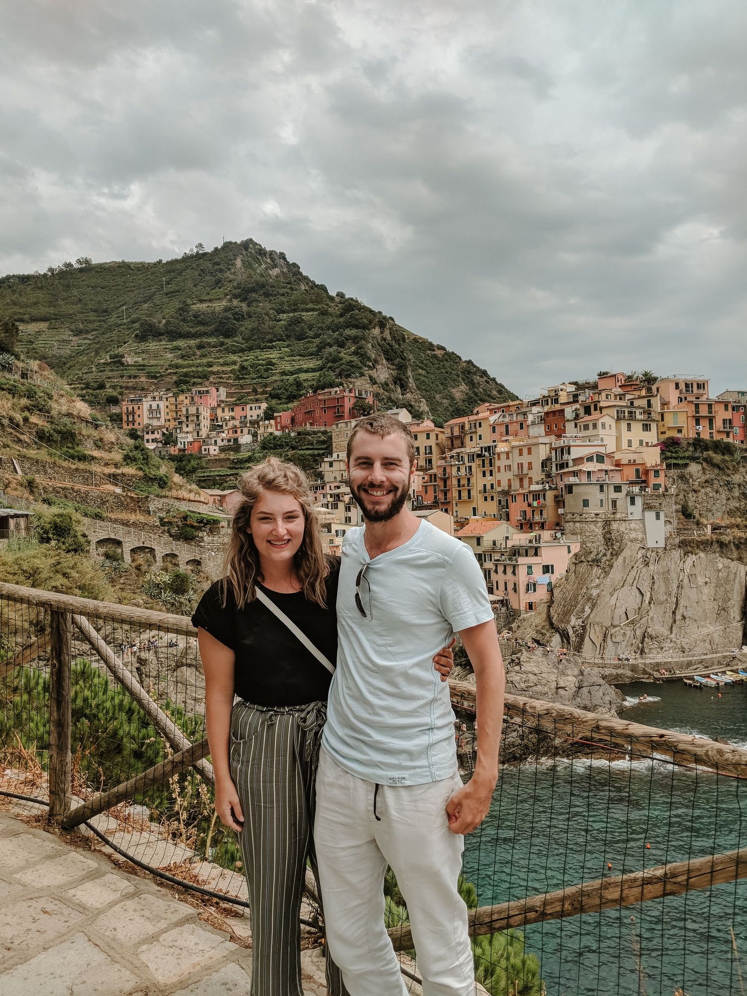 A couple poses in front of the colorful cliffside village of Cinque Terre, Italy with the Mediterranean Sea below.