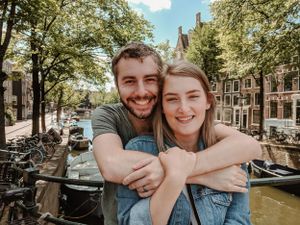 A couple embraces on a sunny day along an Amsterdam canal with boats and historic buildings in the background.