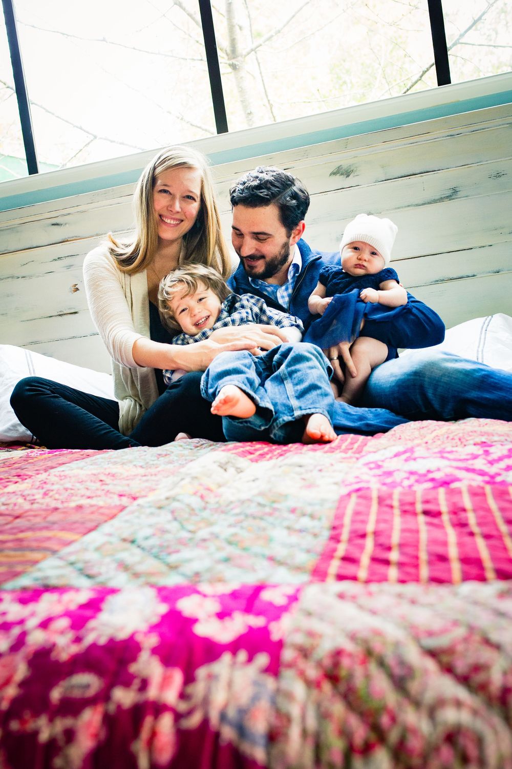 Happy family of four sitting on a colorful patchwork quilt bed, parents smiling with toddler and baby indoors.