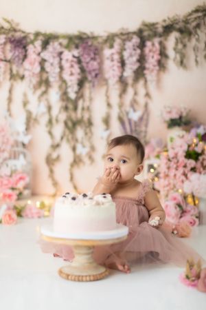 Baby in mauve dress reaching for white birthday cake against floral backdrop.