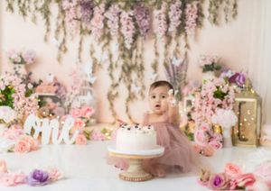 White cake on pedestal with floral decorations and hanging greenery in soft pink tones.