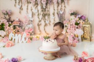 Baby crawling on white floor with pink floral backdrop and cake display for first birthday photo shoot.