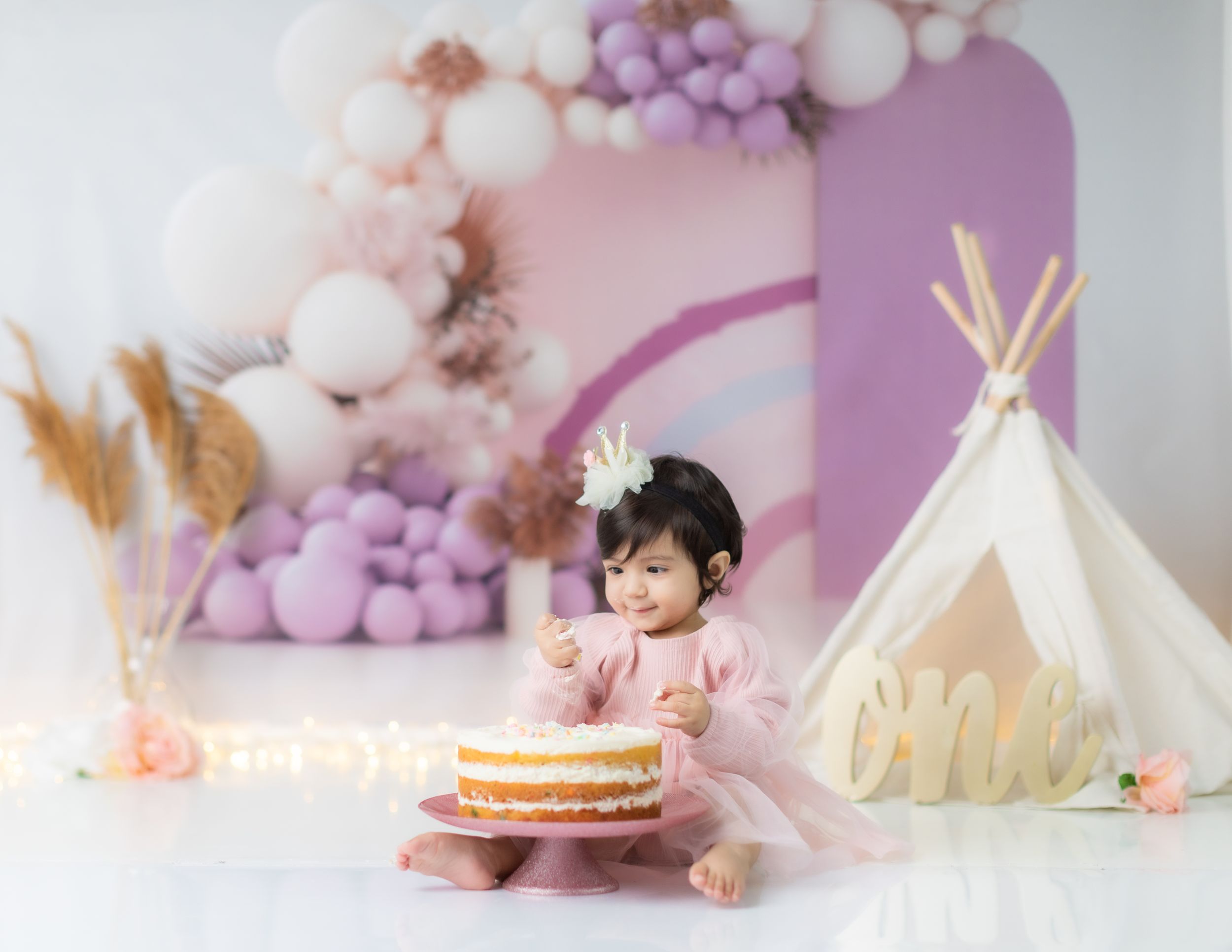 Whimsical birthday setup featuring white teepee and layered cake with purple balloon garland.
