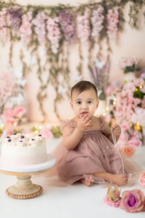 Baby in dusty rose outfit playing near white cake with pink floral backdrop.