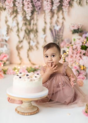 Baby in pink dress lying on floor near birthday cake with floral decorations.