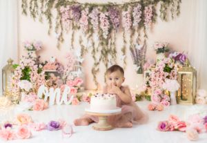 Baby in dusty rose dress sits among pink floral decorations and hanging greenery for cake smash photo session.