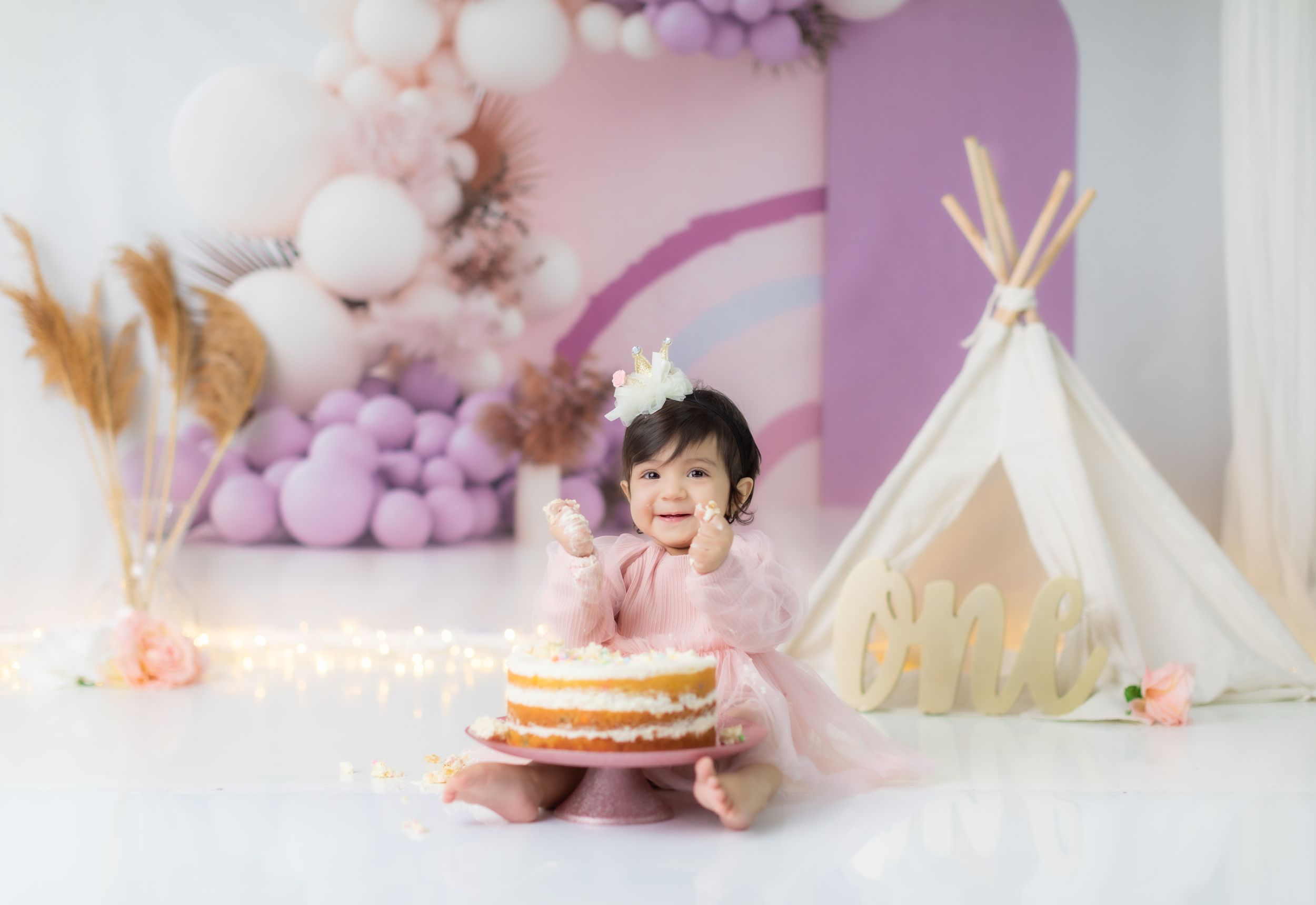 Elegant birthday photoshoot setup with teepee and pink balloons in soft lighting.