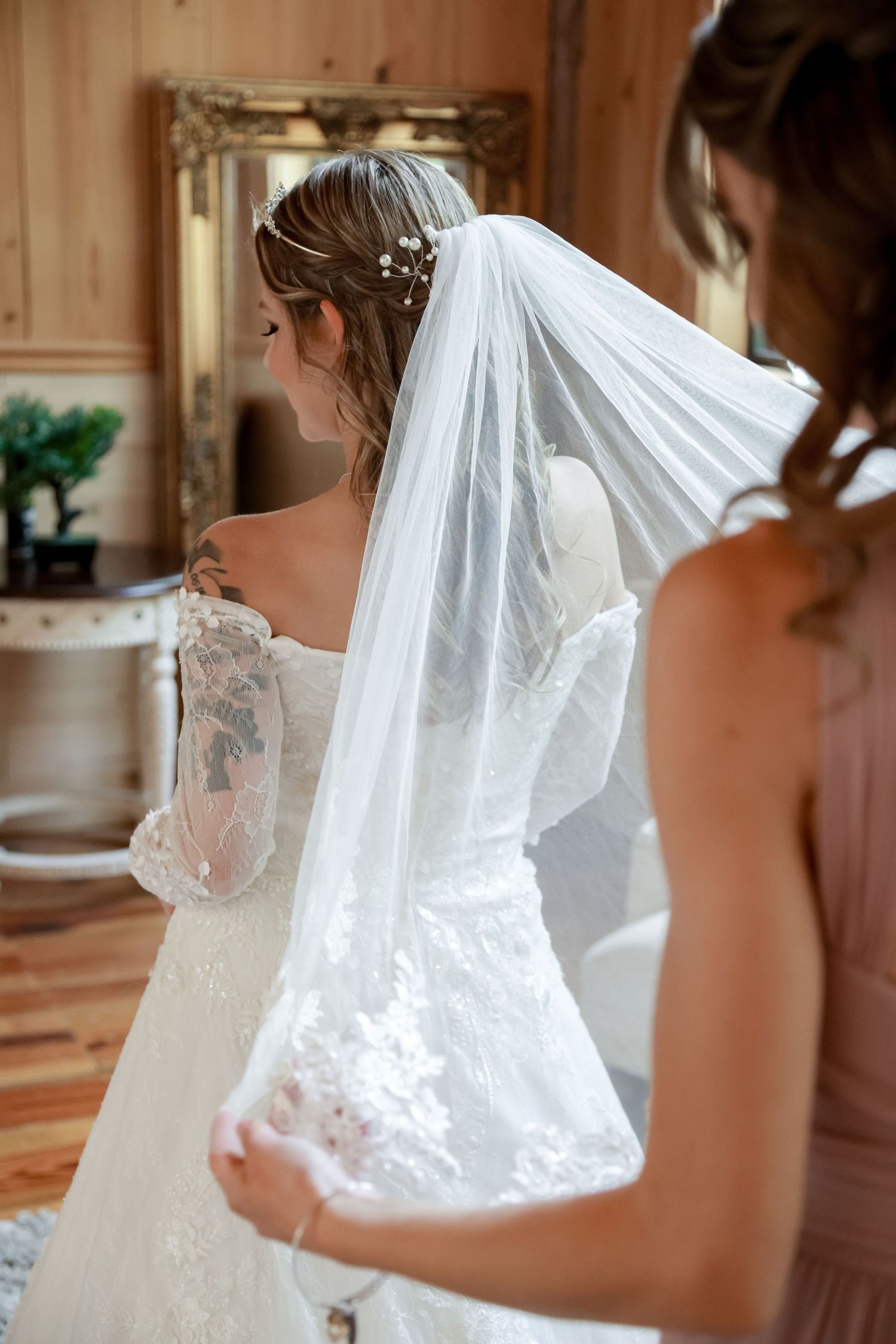 A bride in an elegant lace wedding dress with a flowing veil is photographed from behind in a rustic wooden setting.