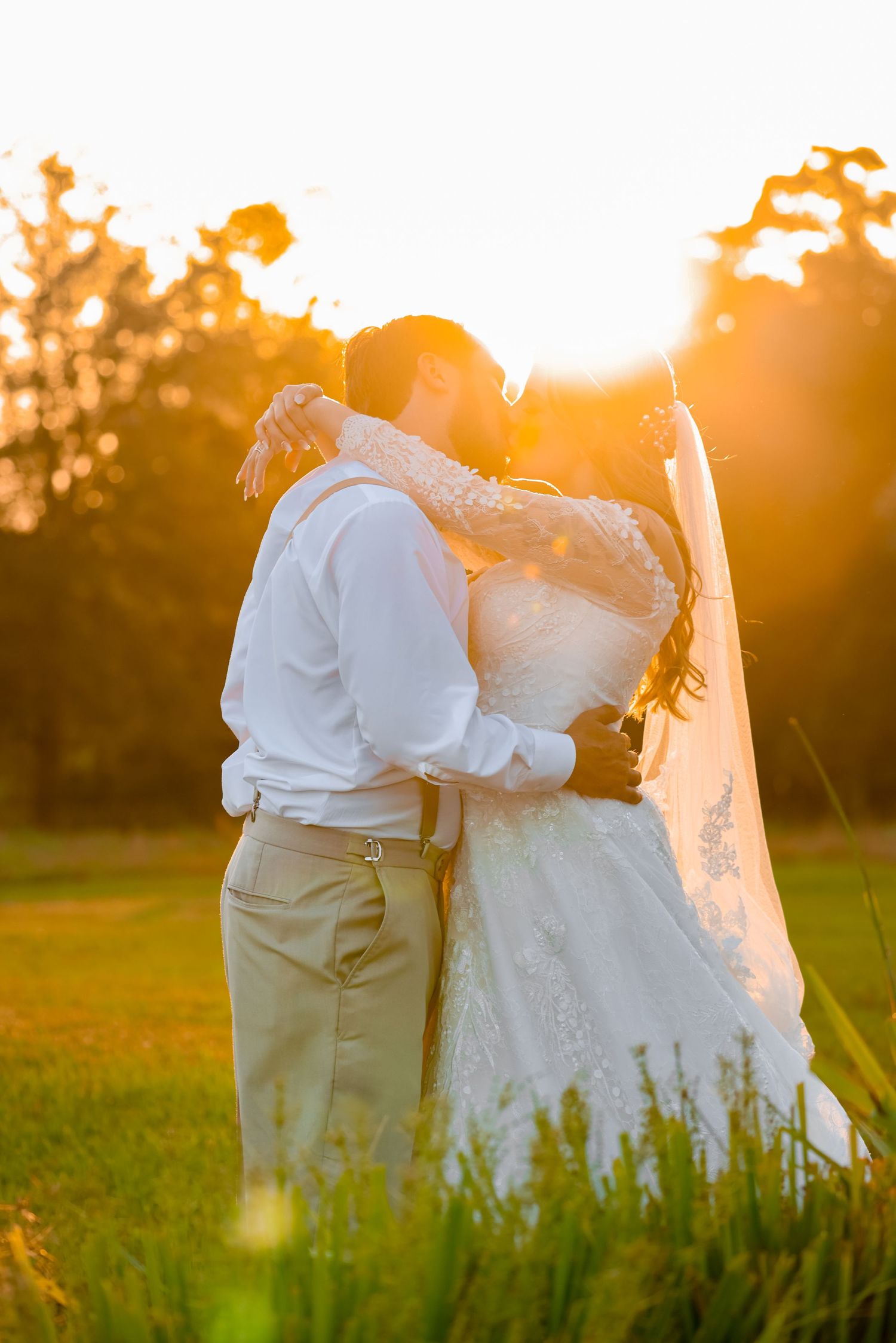 Silhouette of newlyweds embracing during golden hour in a field with flowing veil.