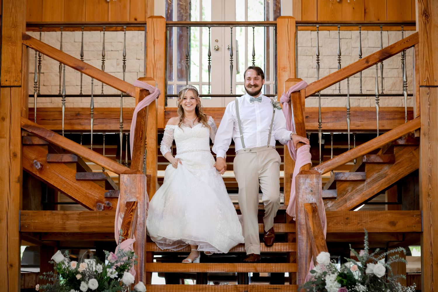 A newlywed couple poses on rustic wooden stairs decorated with white floral arrangements.