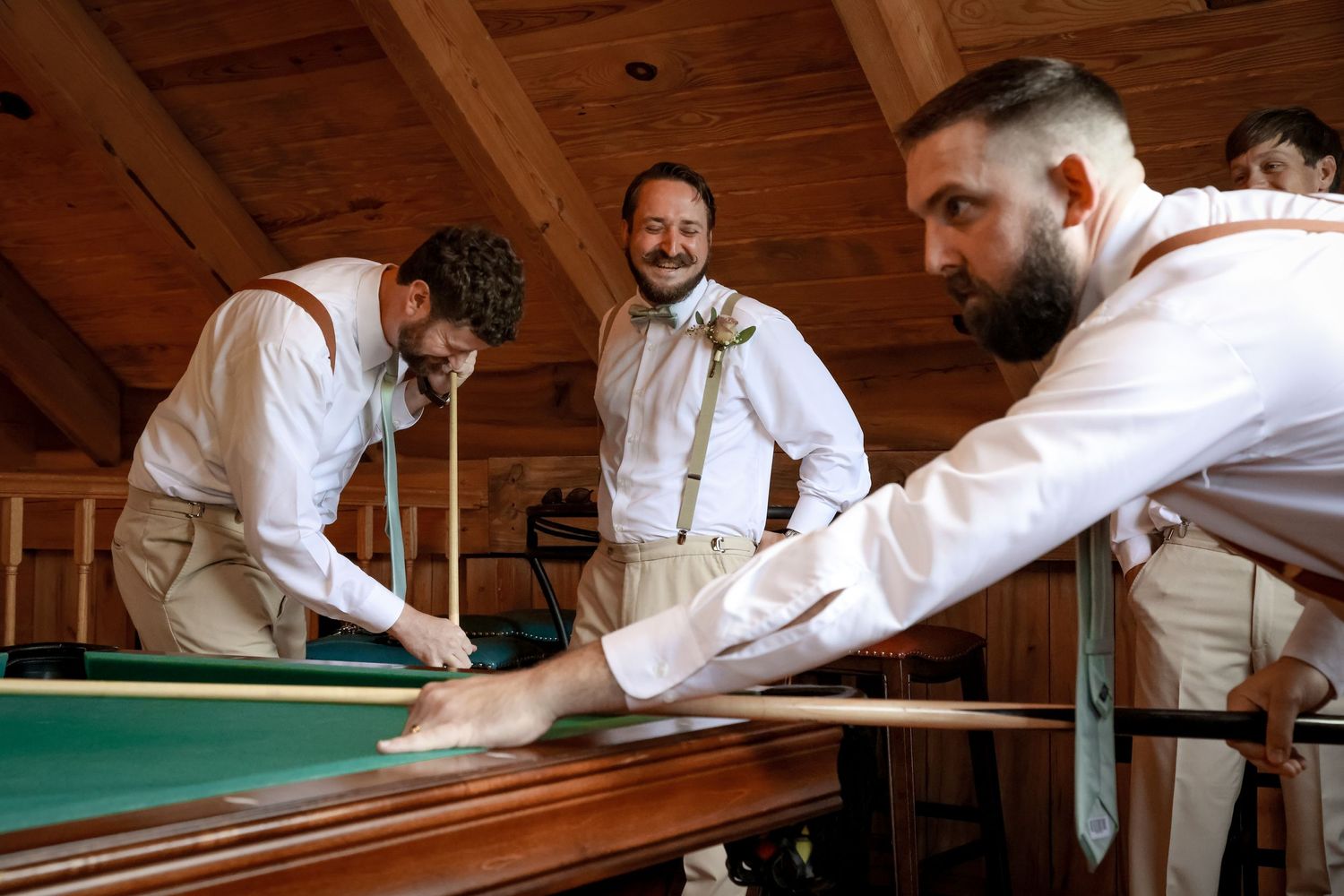 Groomsmen in white shirts and suspenders playing pool during wedding preparations.