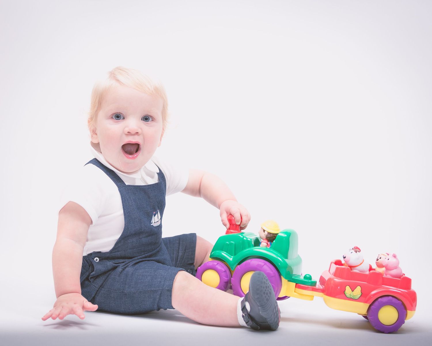 A toddler boy sits against a white backdrop while playing with a toy tractor. He looks at the camera, laughing.