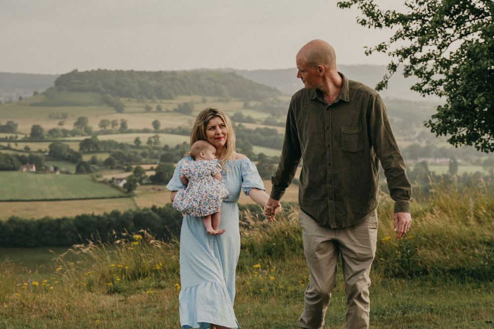 new parents walk with their newborn towards the camera with lovely views of the cotswolds behind them