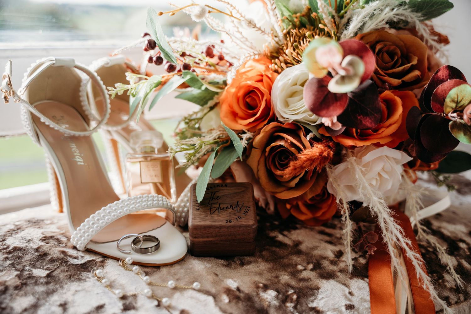 Wedding details with bridal shoes and orange rose bouquet on textured surface.