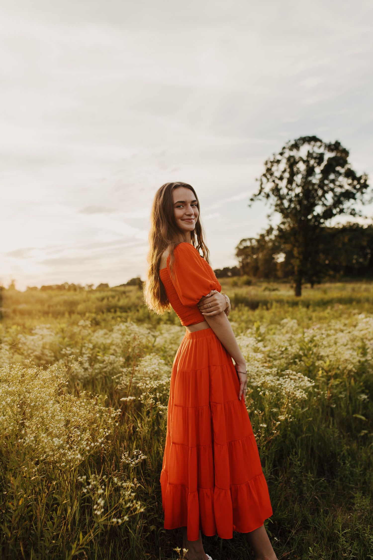 Woman in flowing orange dress poses in golden wildflower field at sunset.