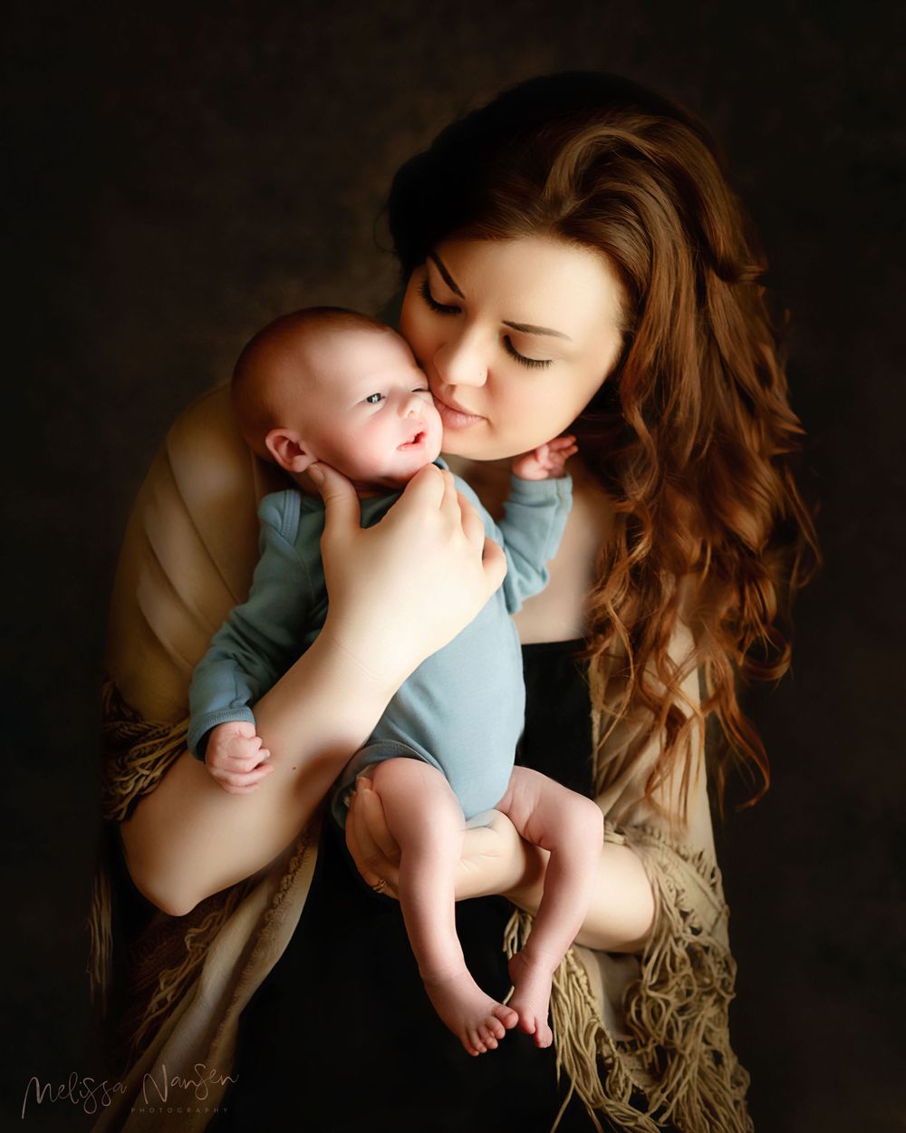 Mother in dark top tenderly kisses baby in blue onesie in emotional portrait.