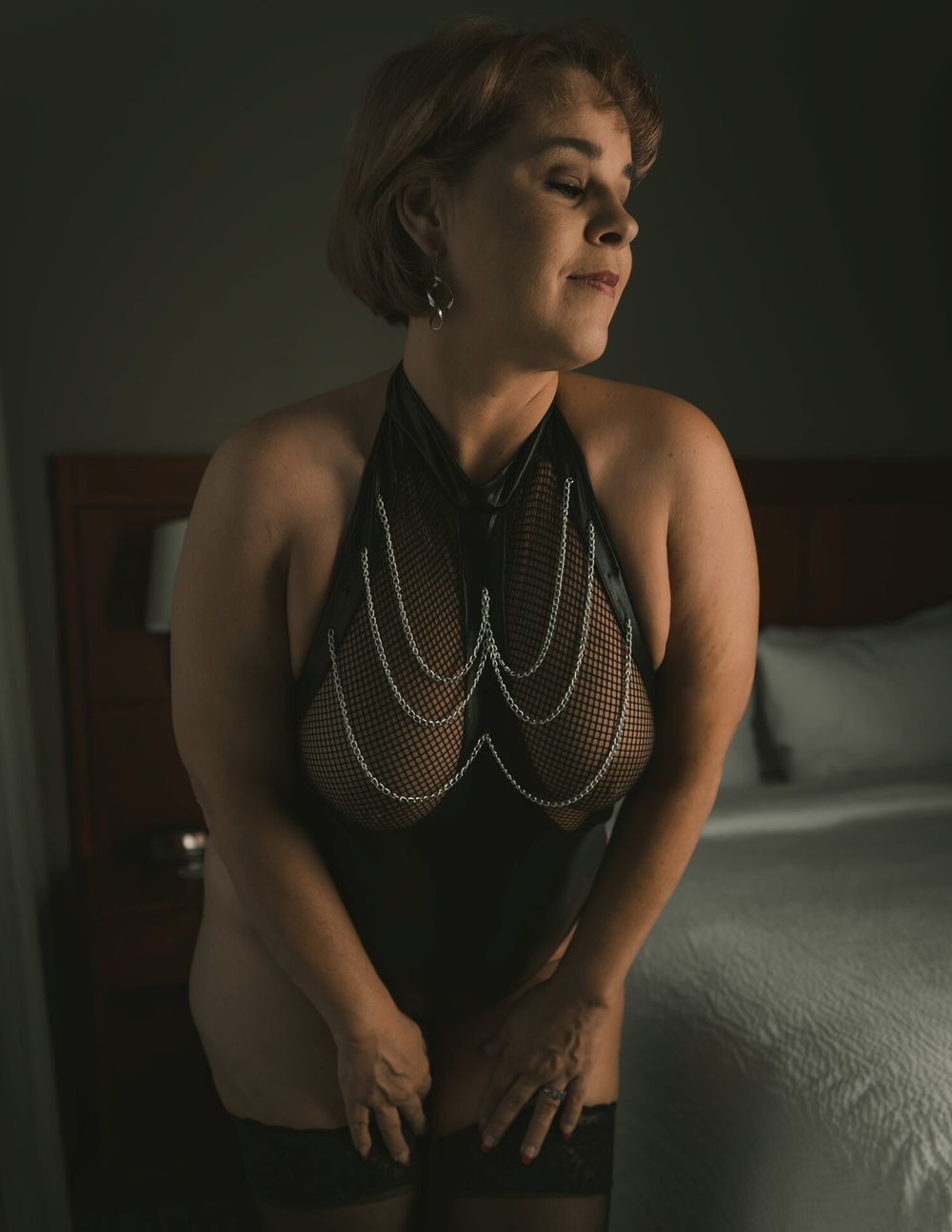 Dark moody boudoir photo featuring a model in a black halter top with draped silver chains in bedroom setting.