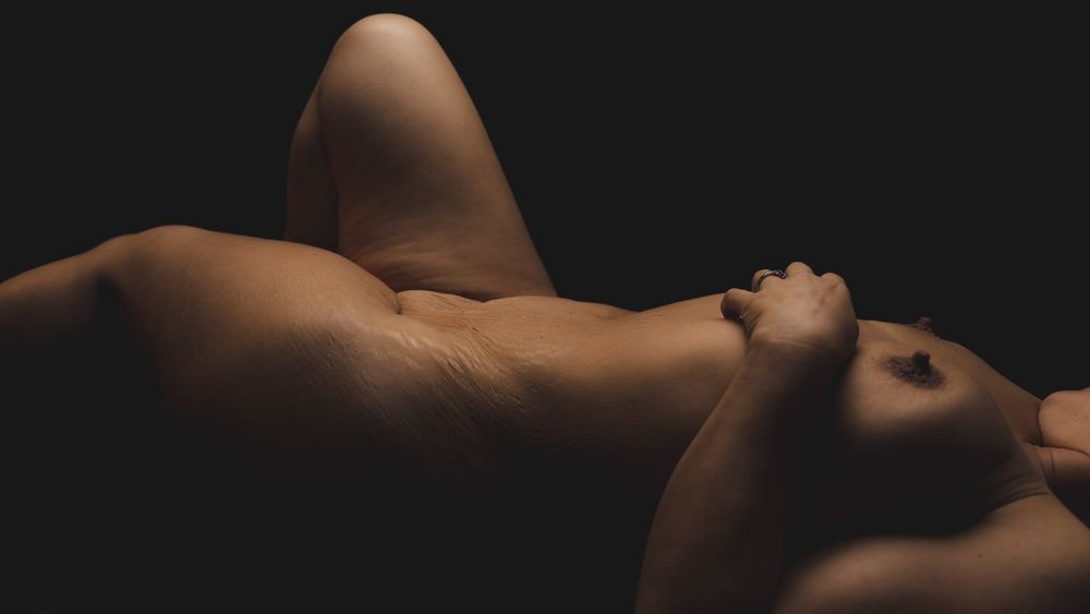 A minimalist artistic nude figure study in chiaroscuro lighting against a dark background.