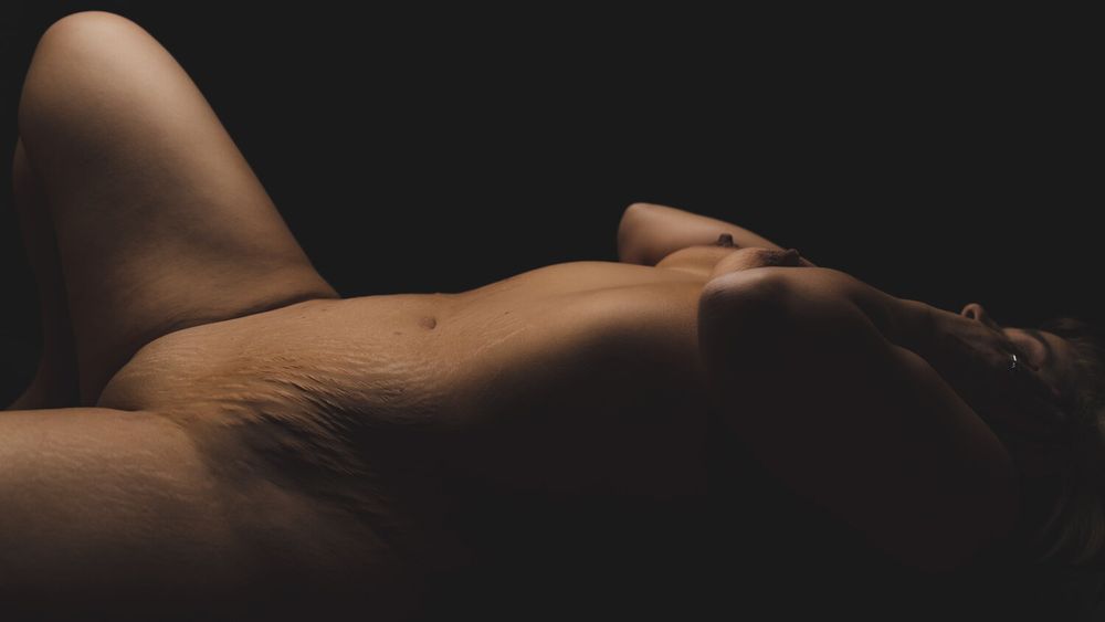 Artistic nude silhouette photographed with dramatic low-key lighting against dark background.