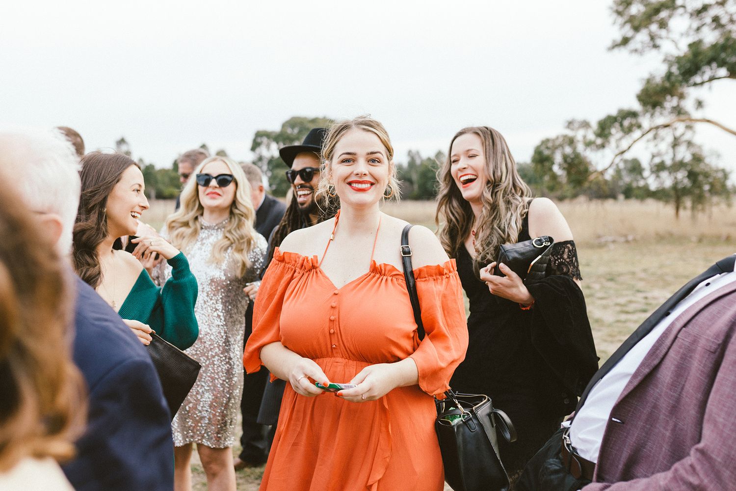 Wedding guests laughing and celebrating outdoors with woman in bright orange dress.
