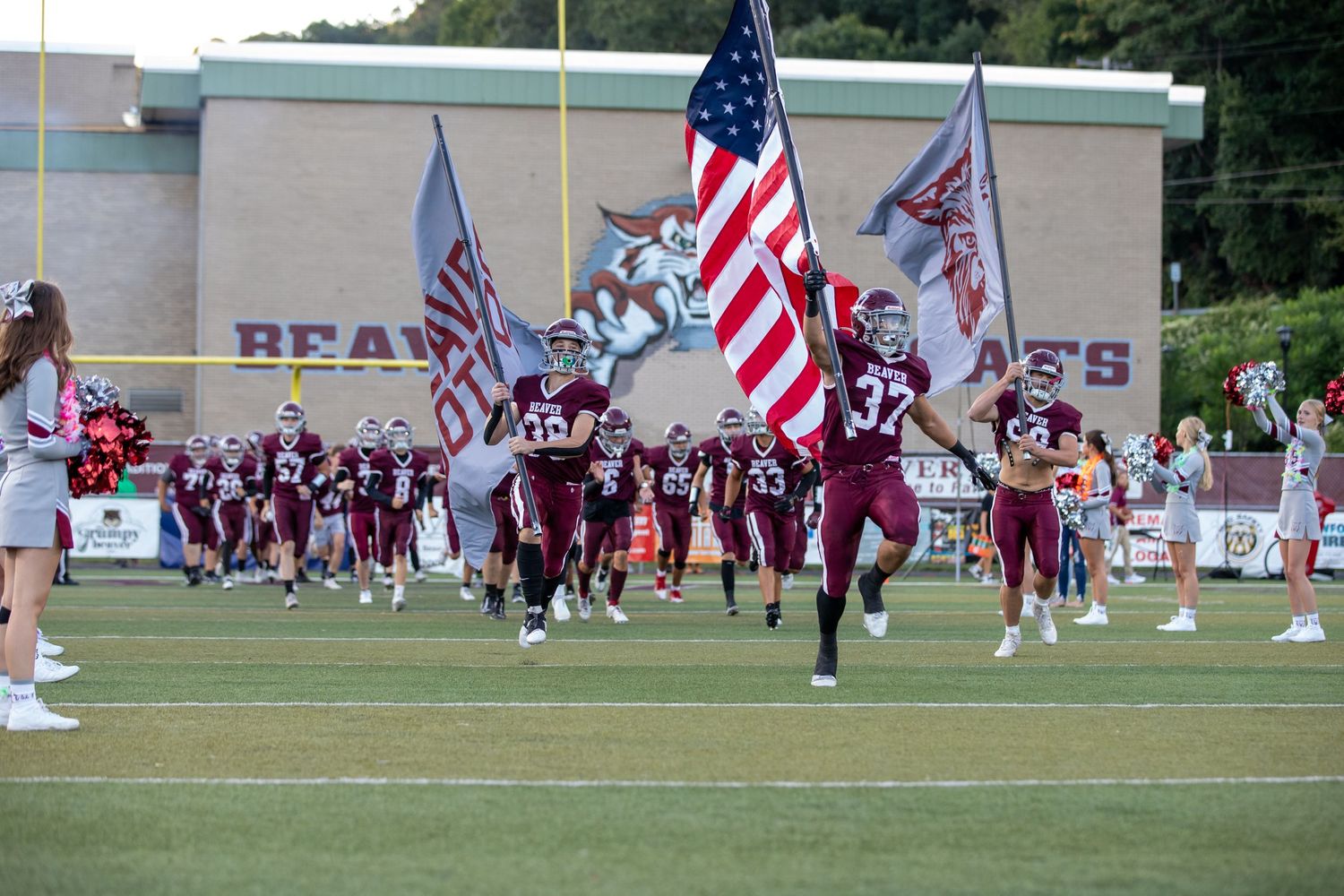 Football players run onto field carrying American flags during pre-game entrance at a high school stadium.