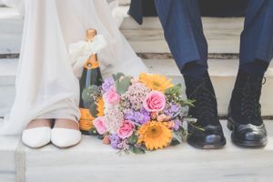 Wedding detail shot showing colorful floral bouquet and formal shoes on stone pavement.