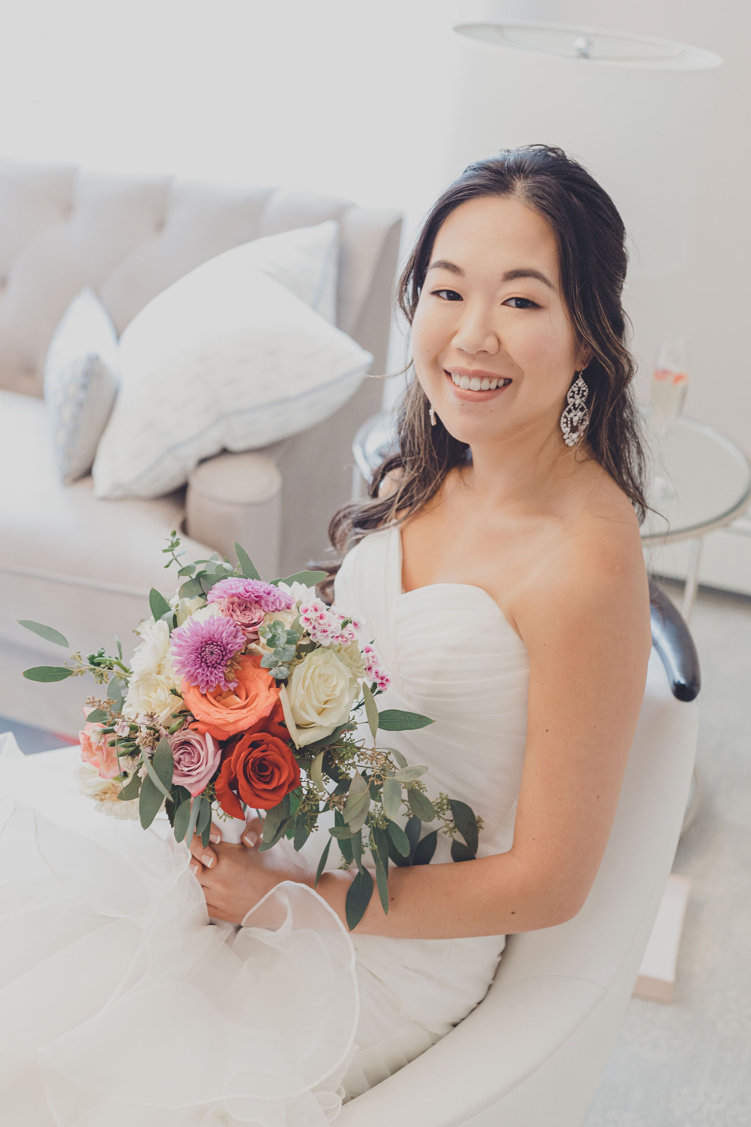 Bride sitting on white couch holding colorful bouquet of roses and dahlias.
