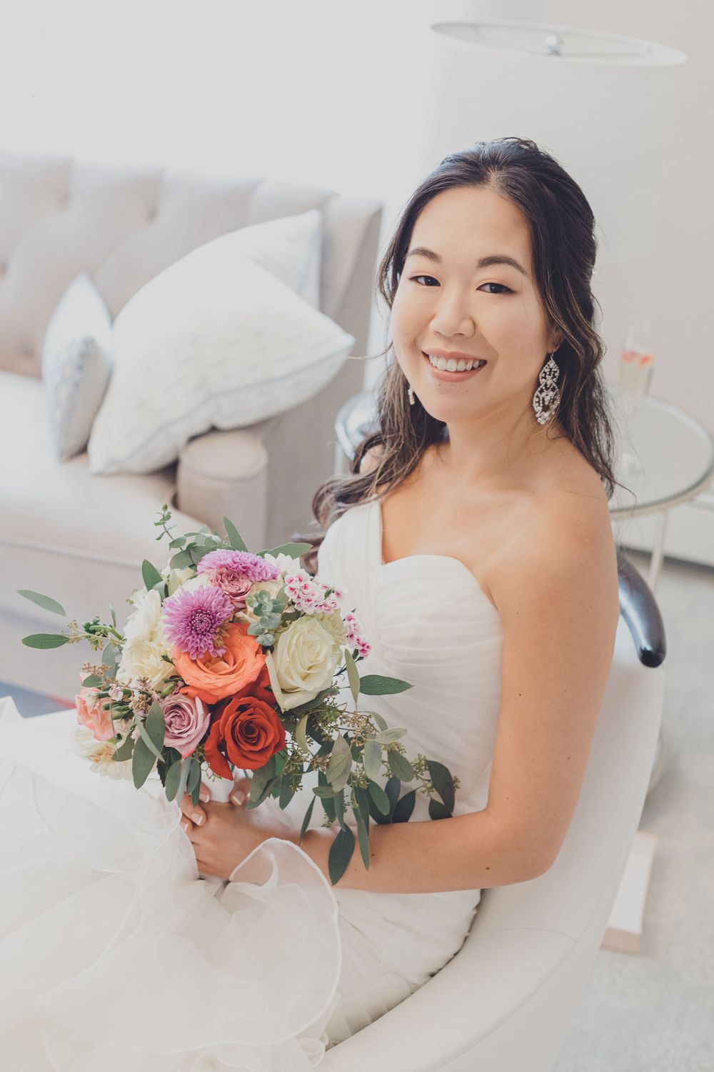 Bride sitting on white couch holding colorful bouquet of roses and dahlias.