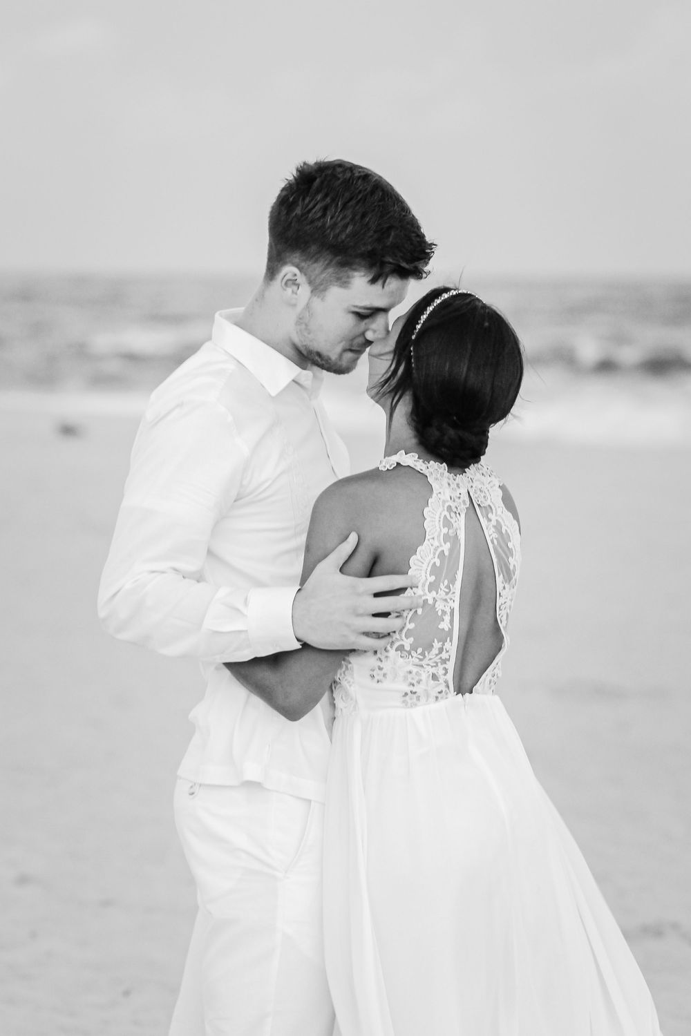A couple in white attire shares romantic moments on a beach in black and white photography.