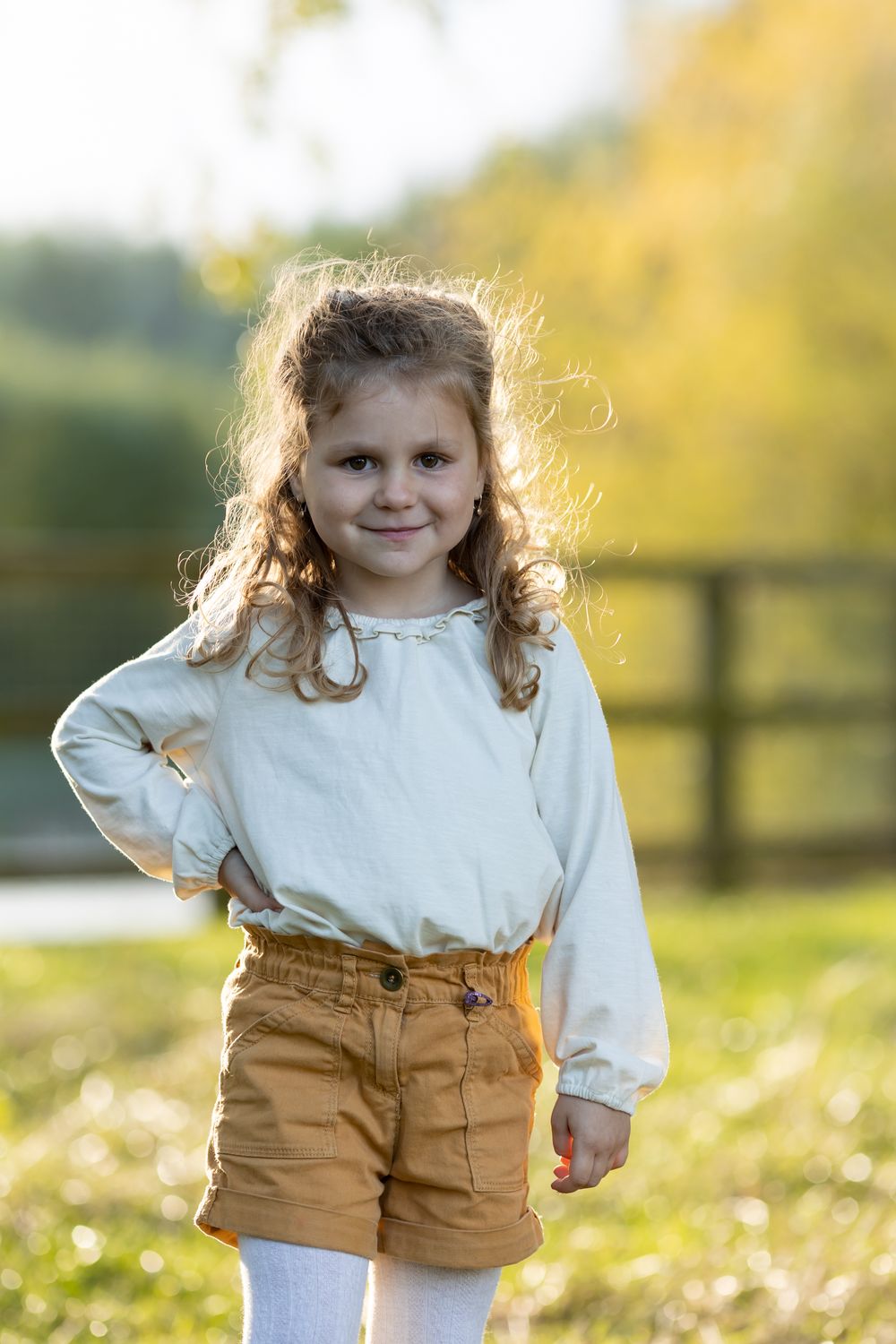 Young child in white shirt and brown shorts poses outdoors with sunlit autumn background.