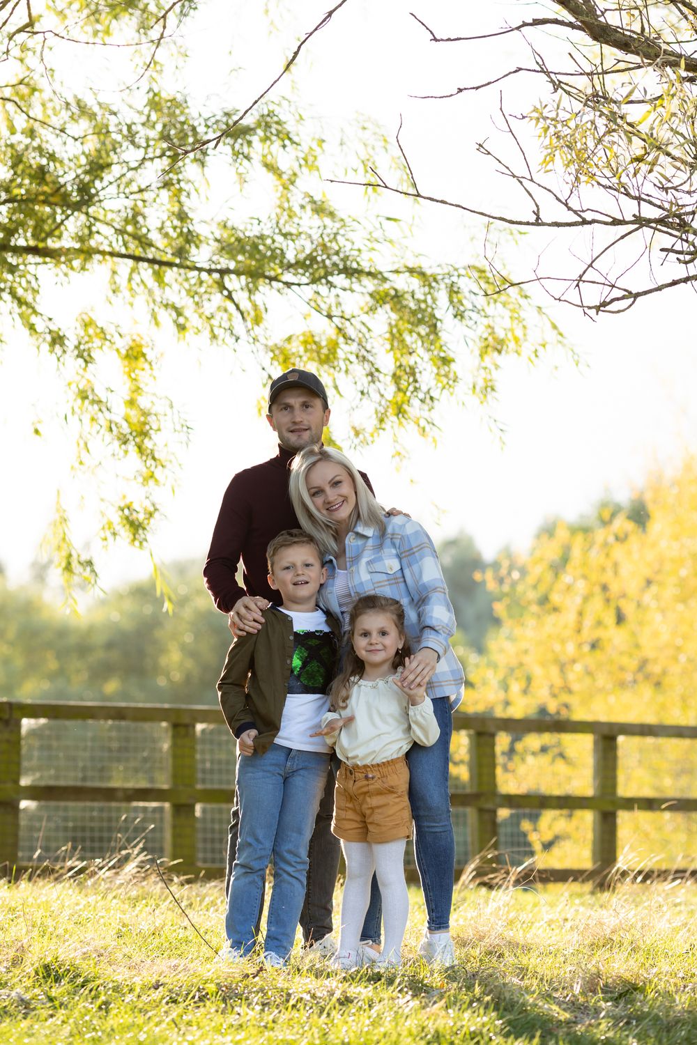 Family standing together in front of a wooden fence and autumn trees during sunset.
