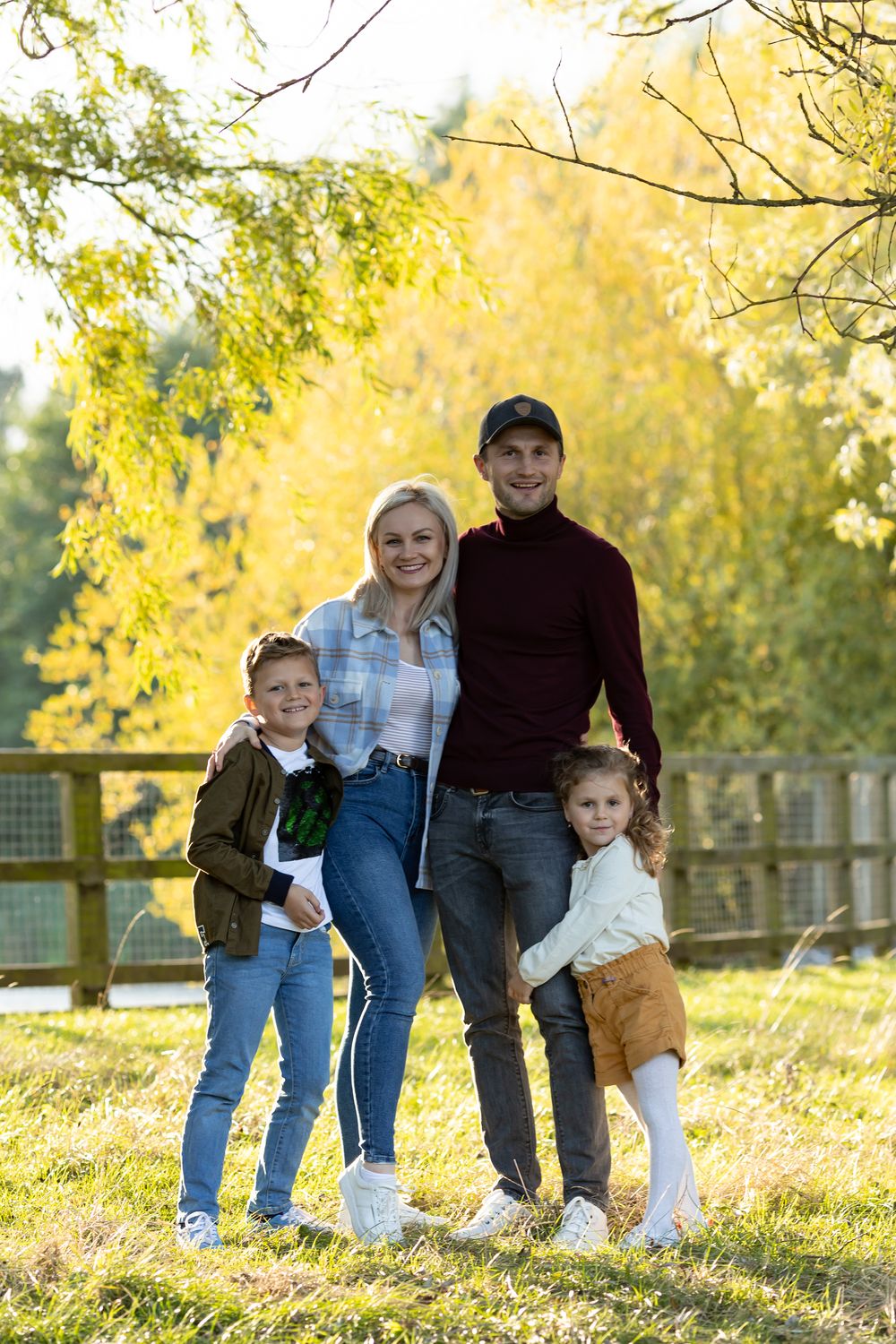 Family portrait outdoors with autumn foliage and wooden fence in background.