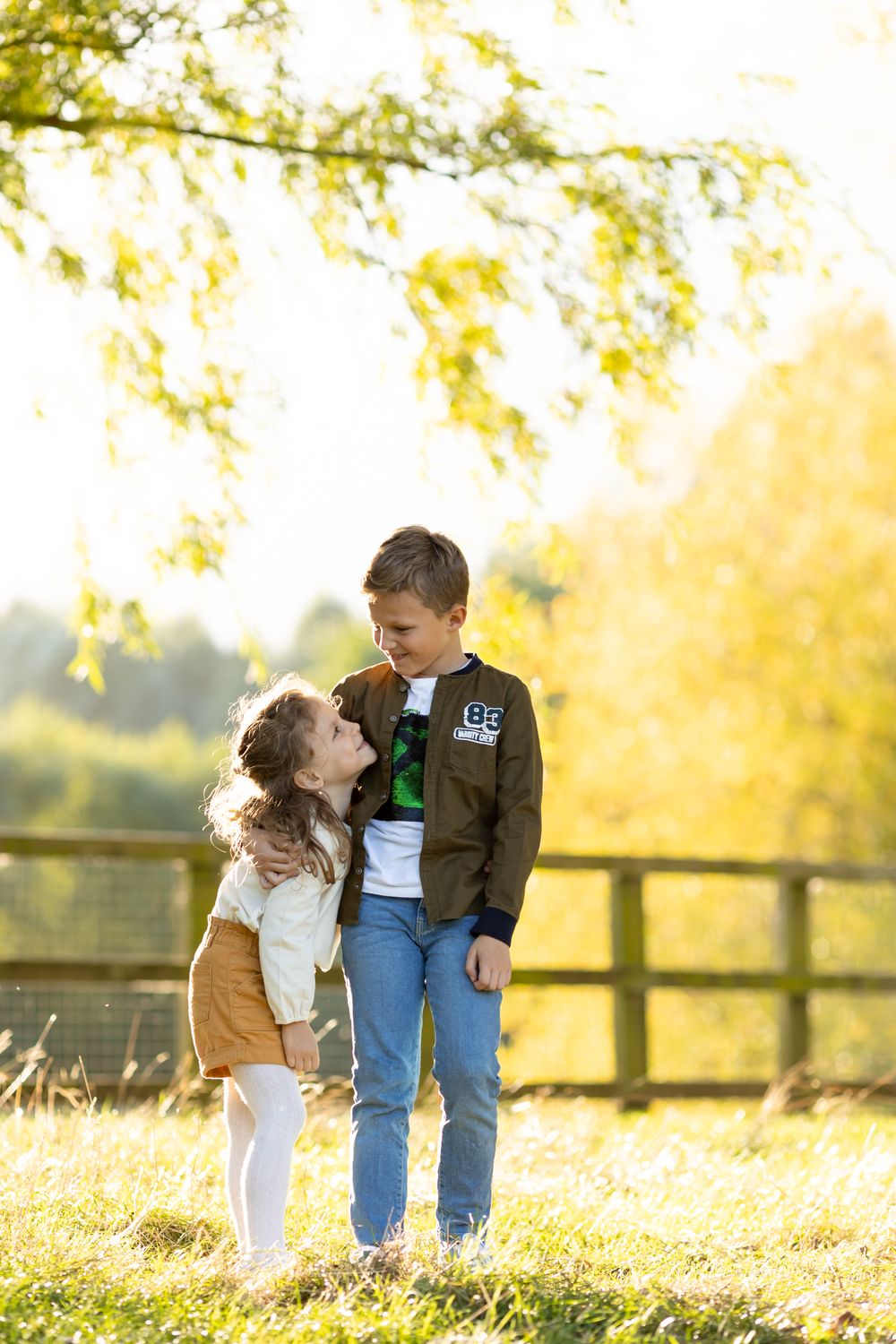 Two children share a sweet moment together in front of a rustic fence during golden hour.