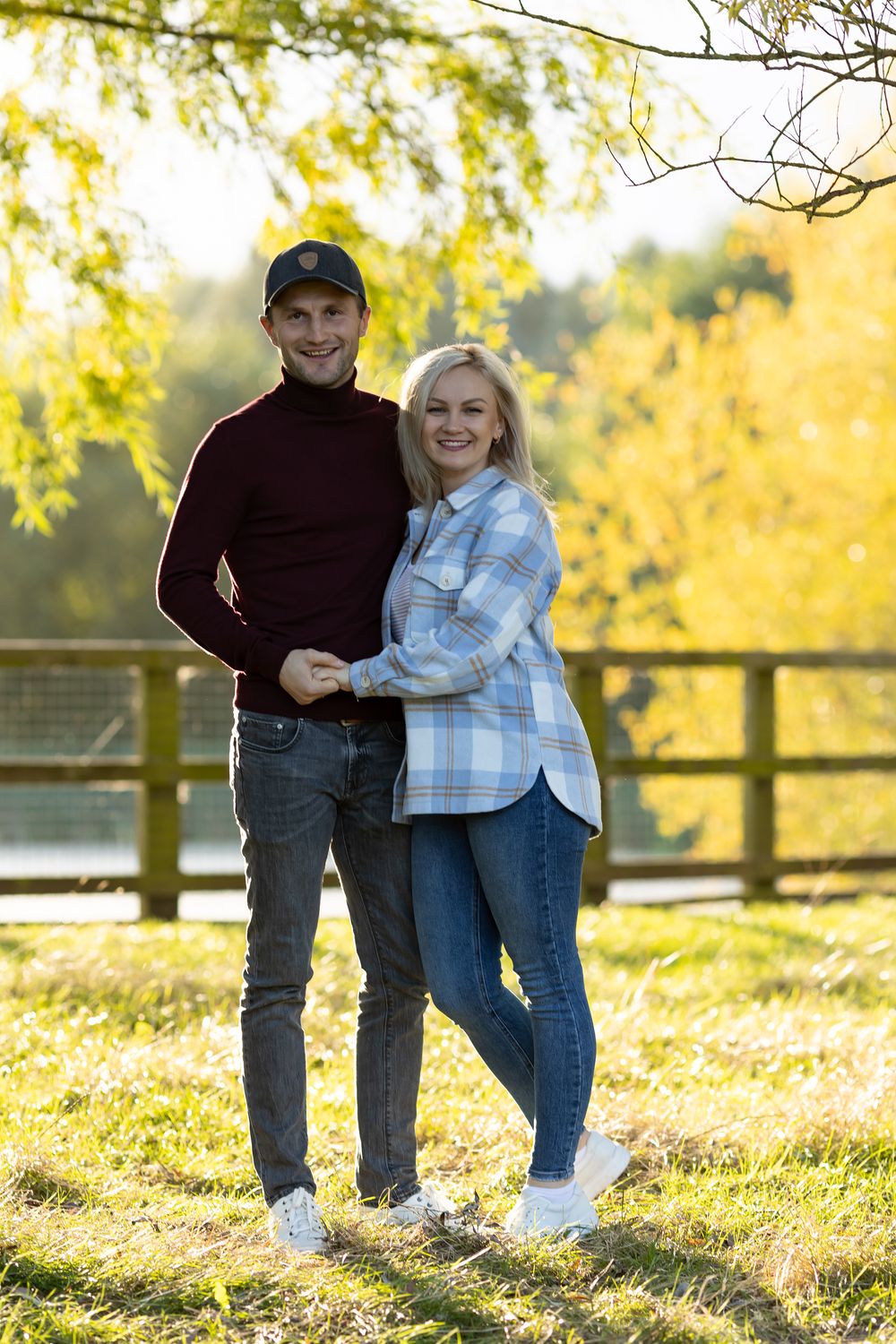 Couple poses together in casual attire against backdrop of golden autumn trees.