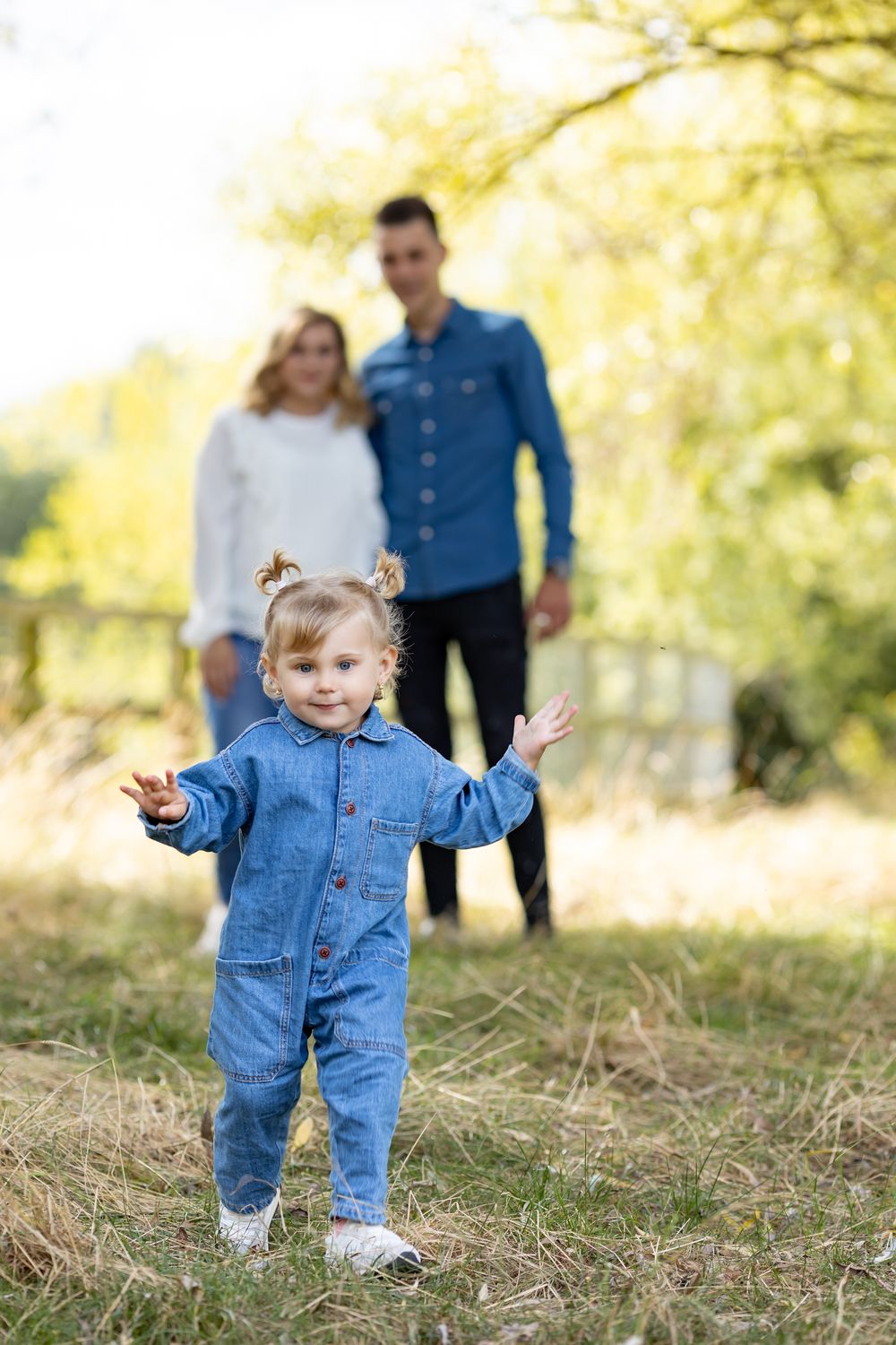Toddler in denim jumpsuit plays in foreground while parents watch from behind.