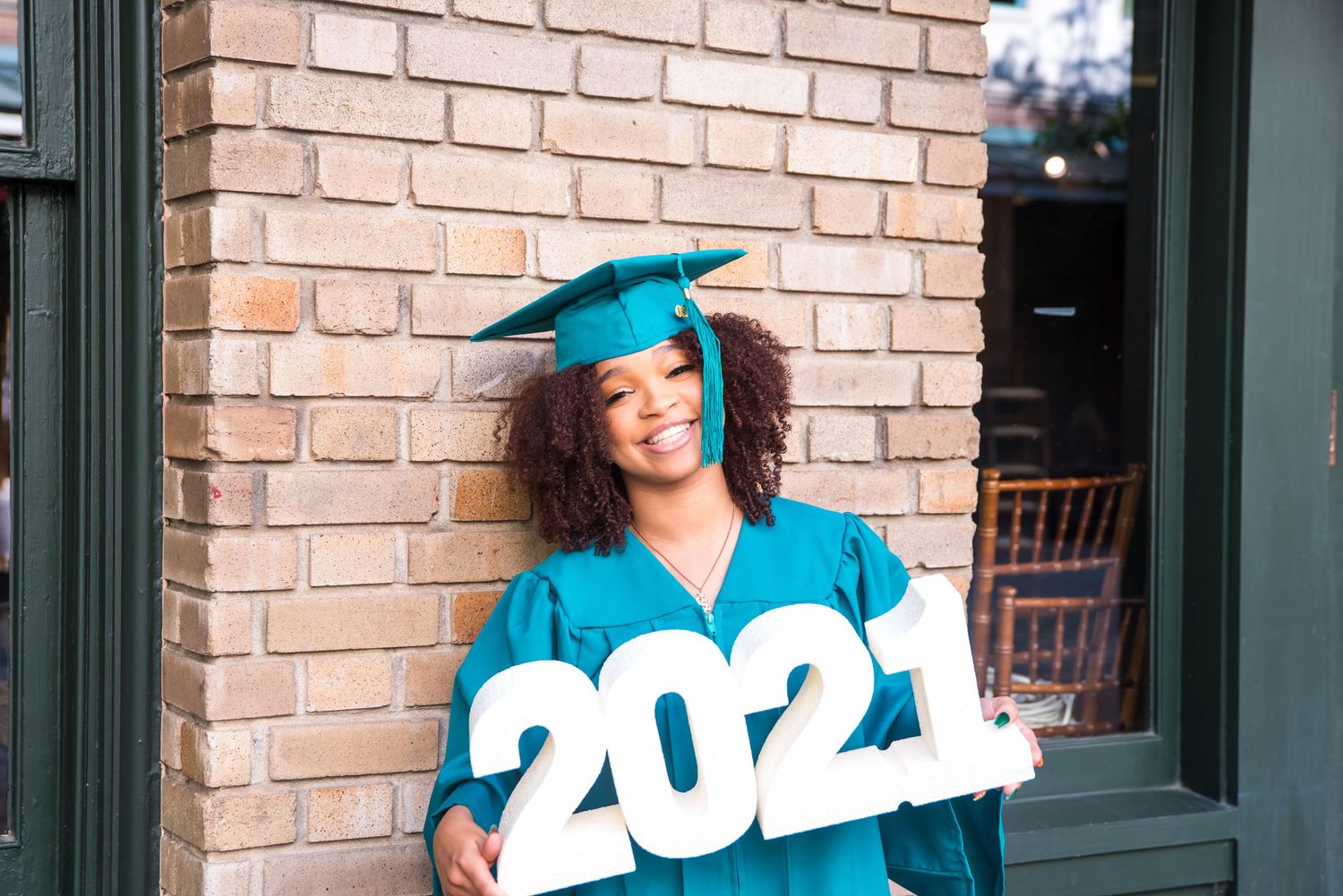 Graduate in teal gown holds 2021 numbers while posing against brick wall.