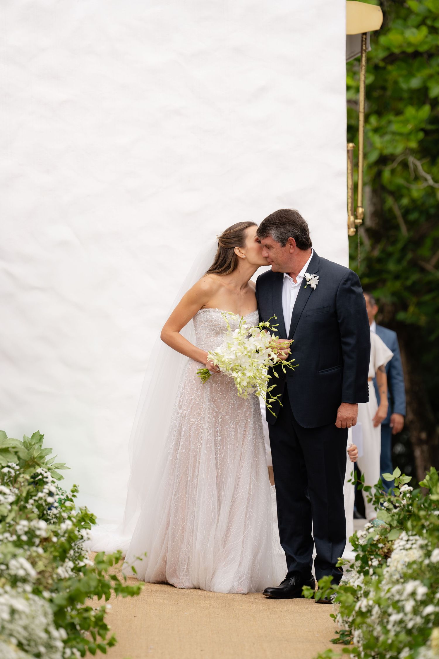 Romantic moment between couple on flower-lined wedding path with white wall backdrop.