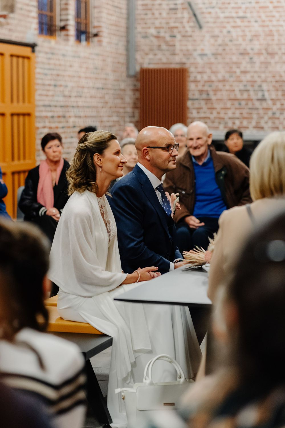 A smiling couple sits side by side during an intimate indoor wedding ceremony with guests in the background.