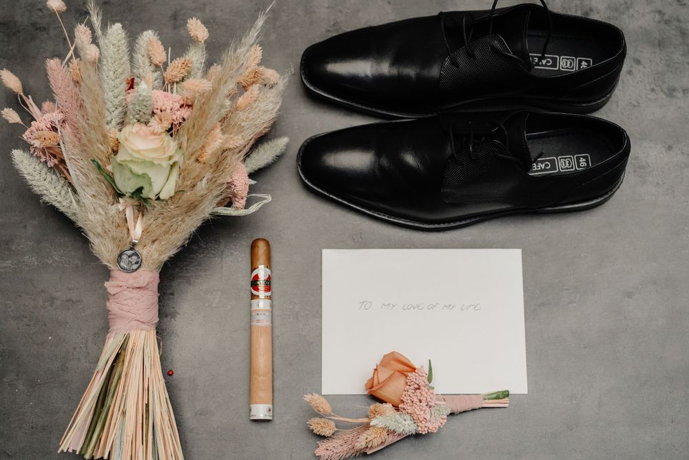 A wedding flatlay with black leather dress shoes, dried floral bouquet, cigar and blank paper note on gray background.