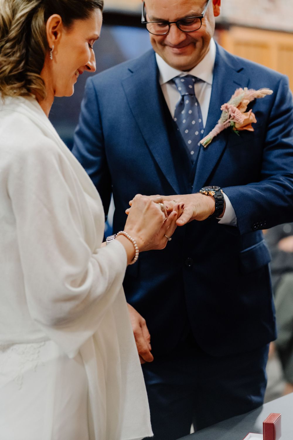 At an intimate wedding ceremony, a couple exchanges rings while the groom wears a navy suit with a pink boutonniere.