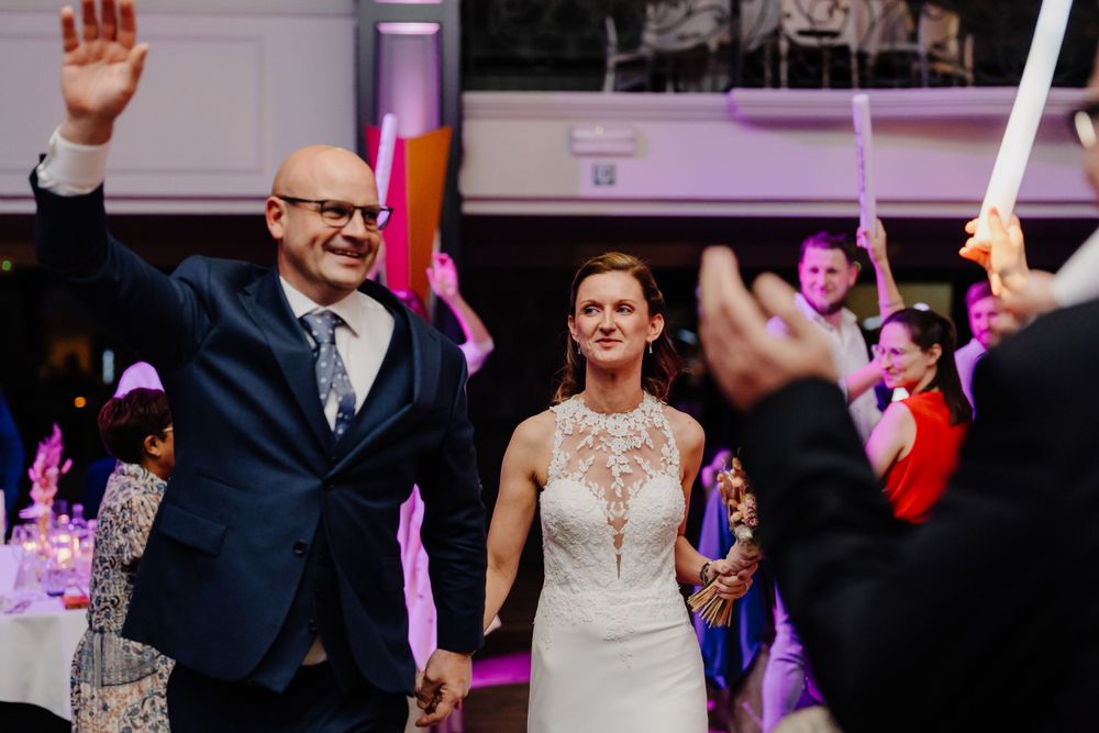 A well-dressed couple celebrates with joy on their wedding day surrounded by dancing guests and pink lighting.