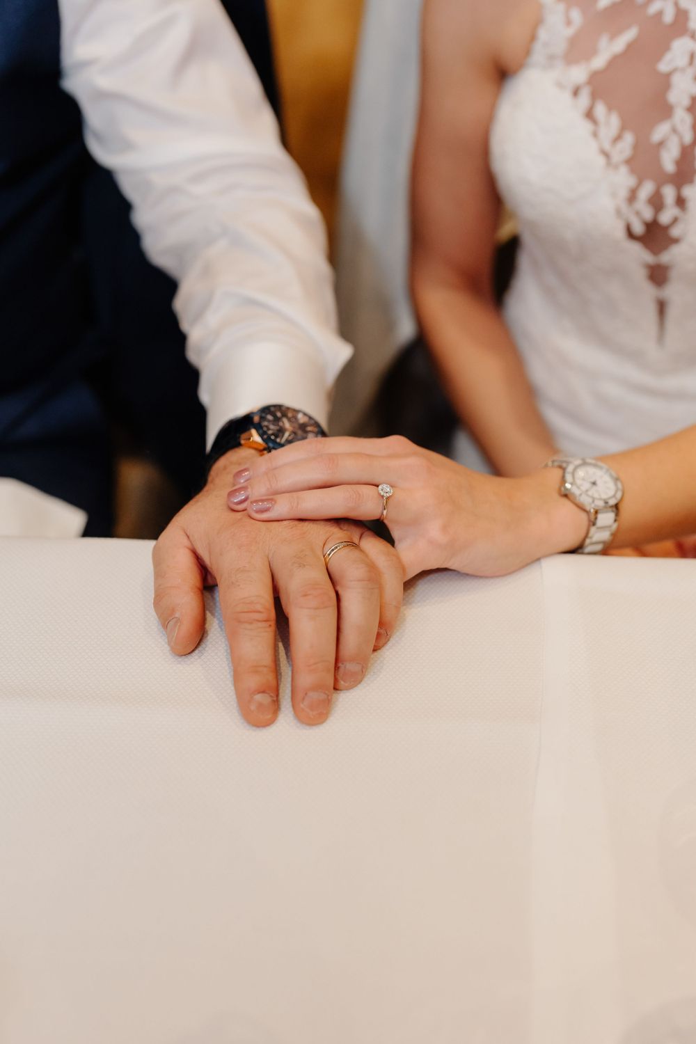 Close-up of newlywed couple's hands with wedding rings, showing wristwatch details on a white tablecloth.