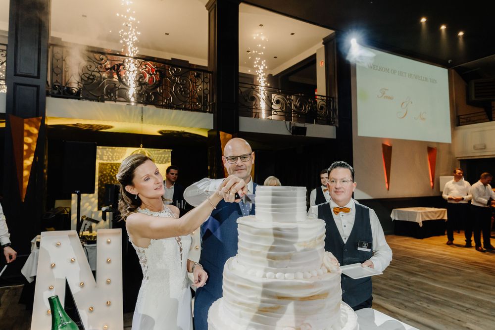 Wedding couple and guest perform a champagne pour during their reception next to a tall white wedding cake.