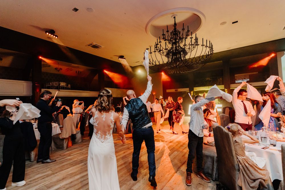 Wedding reception guests dance on a wooden floor under a chandelier with warm lighting and table settings visible.