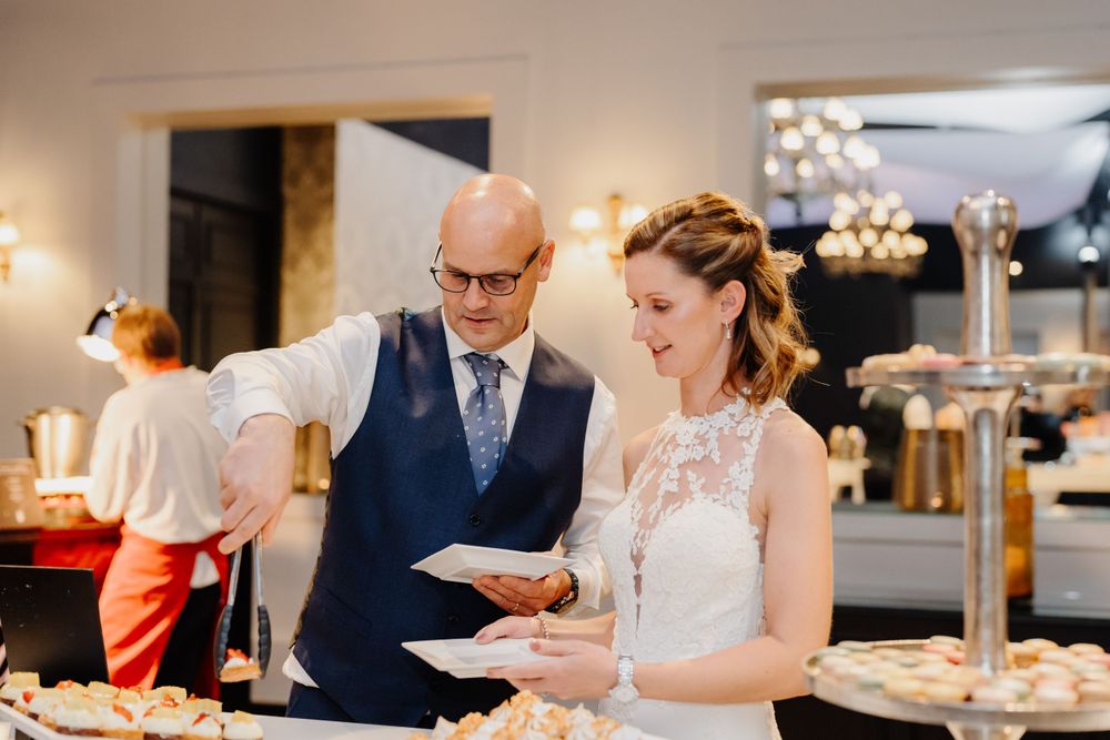 A couple examines desserts at an elegant reception buffet table with warm lighting and formal attire.