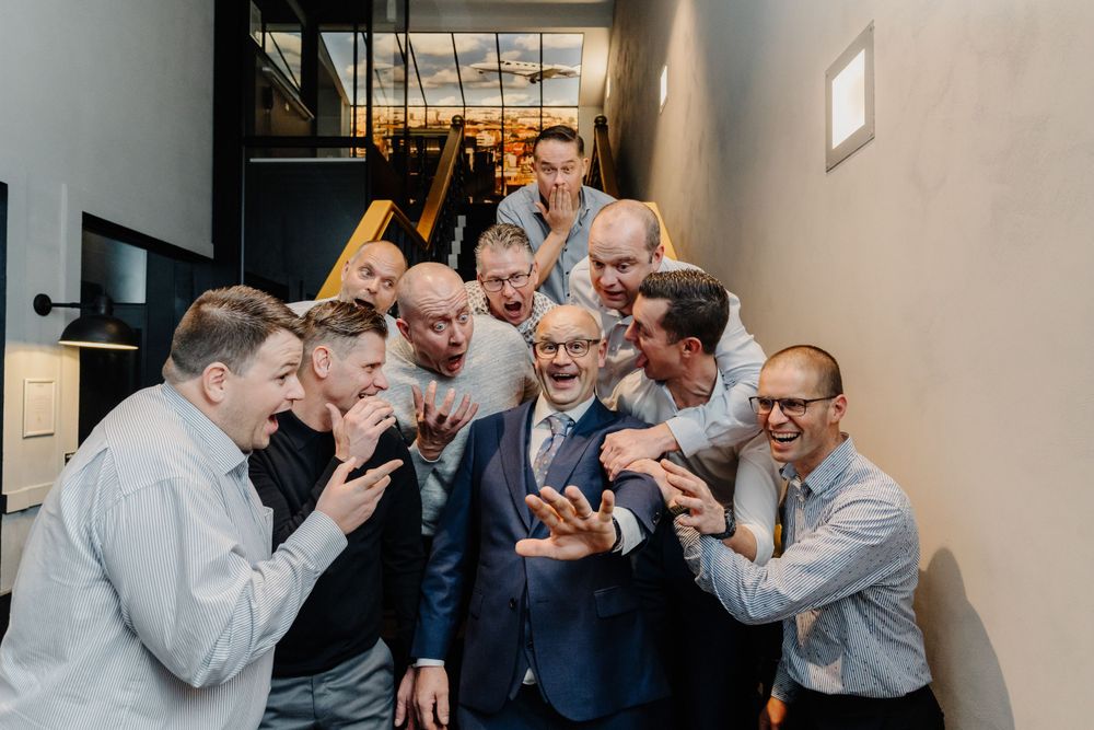 A group of formally dressed men gather in a hallway reacting excitedly while looking at something on a mobile phone.