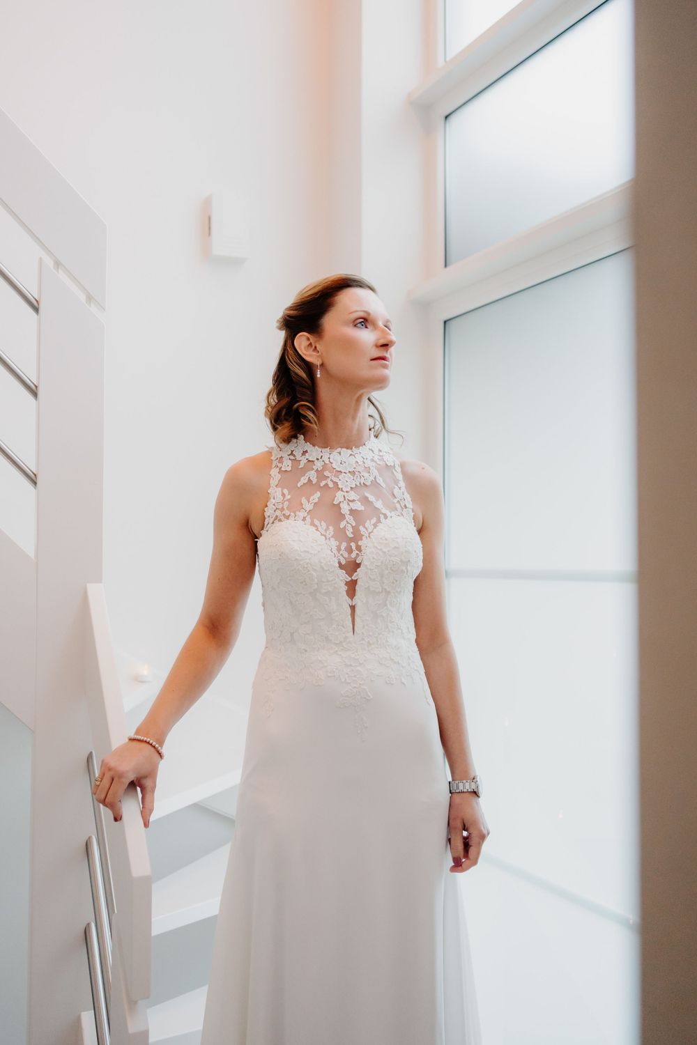 A bride in an elegant white wedding gown with lace bodice and high neckline stands near a bright window.