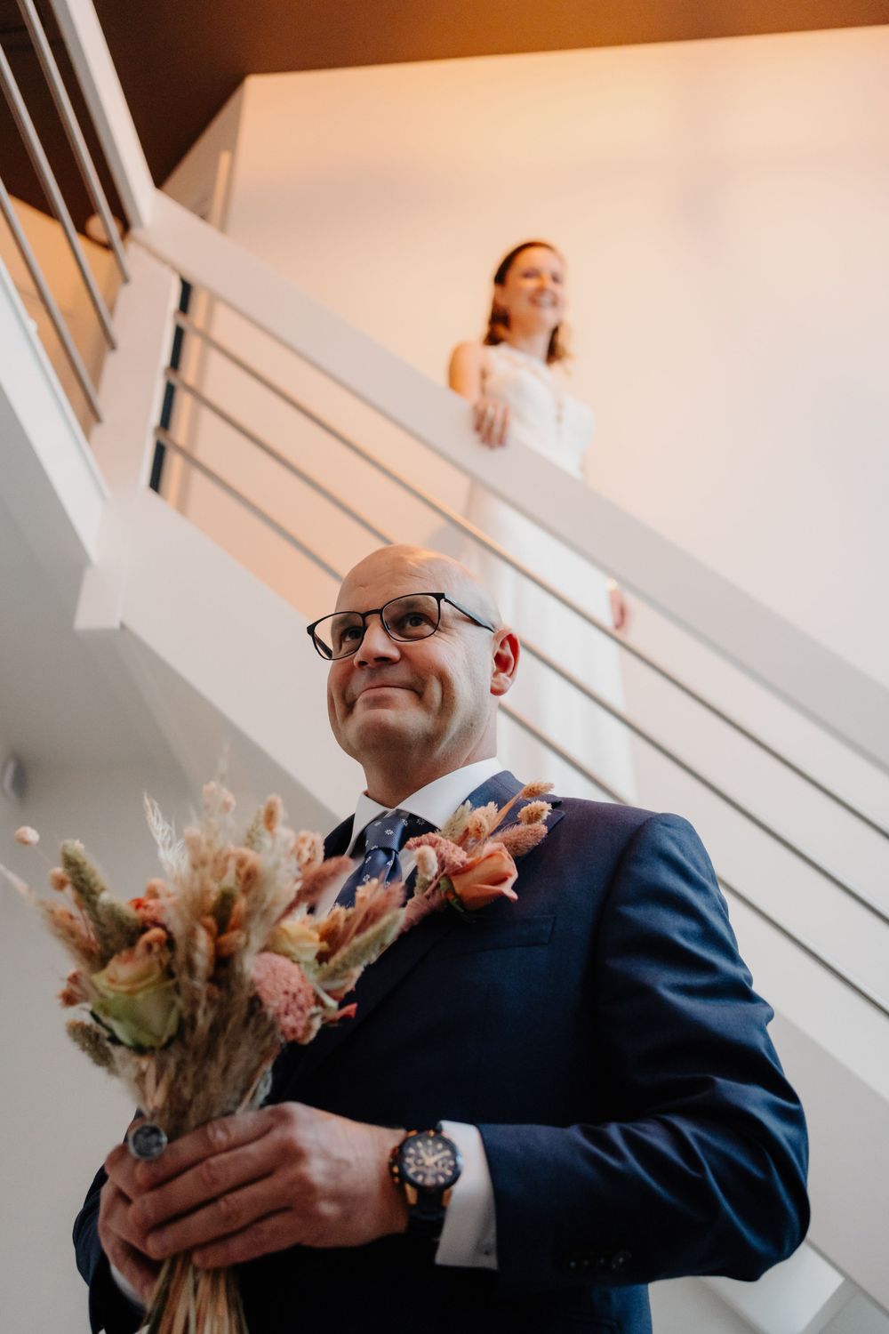 A wedding scene shows someone in a navy suit holding a romantic bouquet of flowers while looking up a white staircase.
