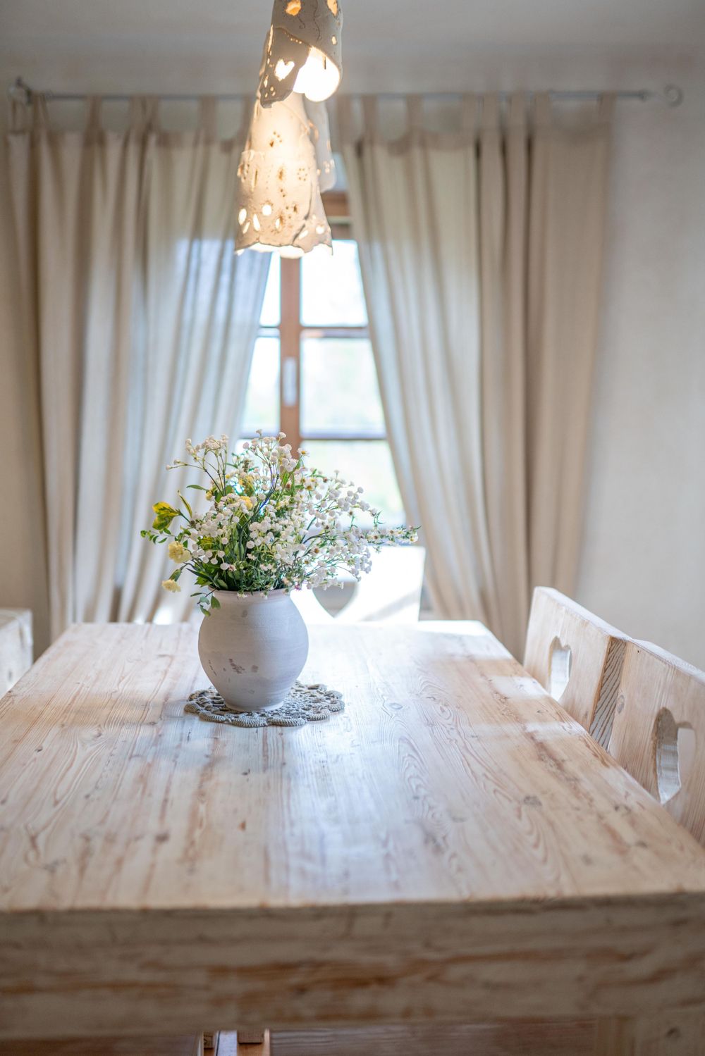 Rustic dining room with white flowers in vase on wooden table near window with flowing curtains.