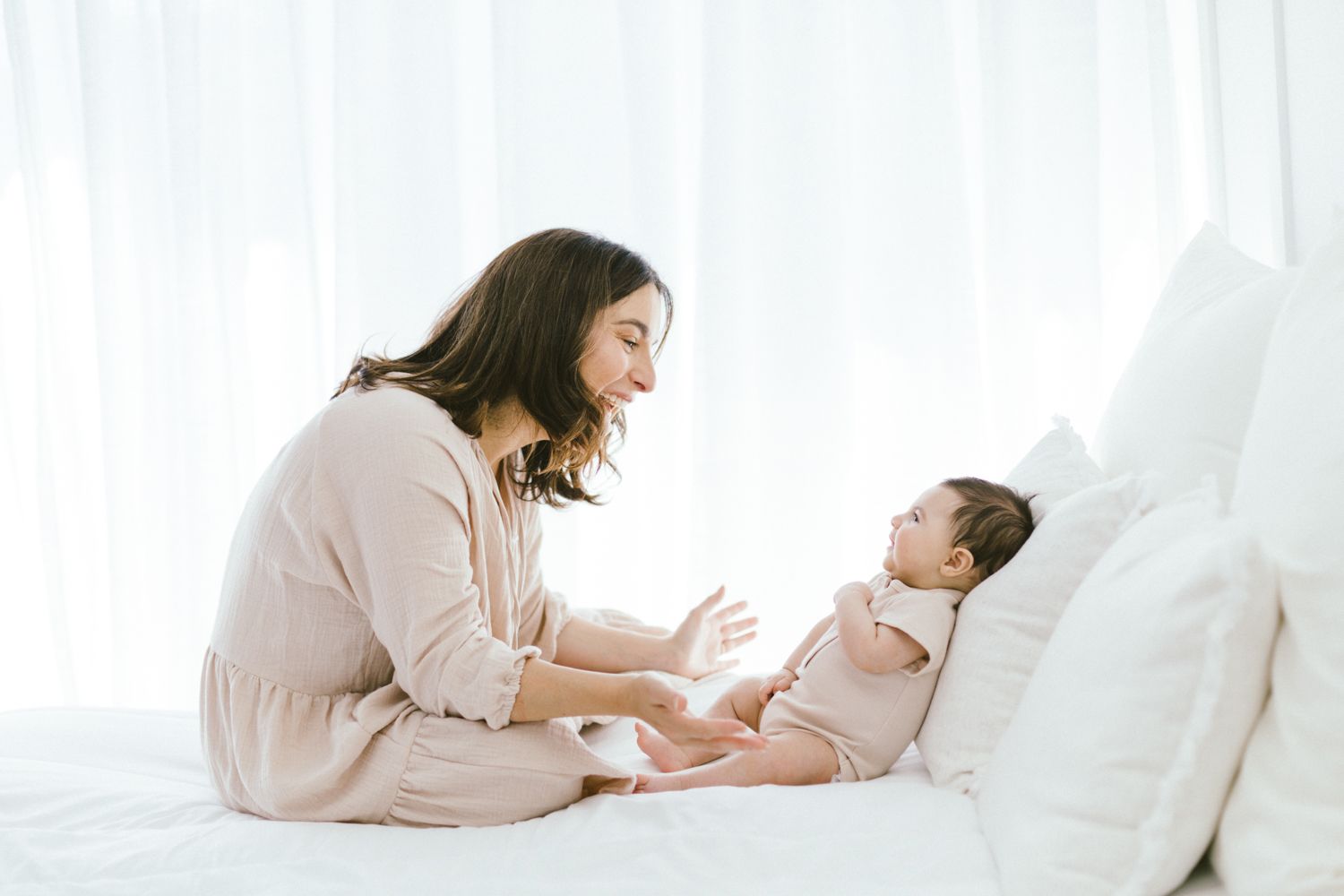 Mother in soft pink clothing snuggles with baby on white bedding in tender moment.